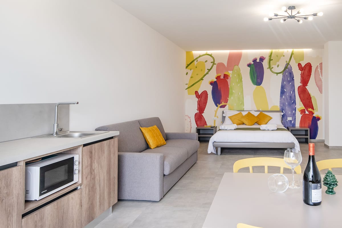 Un espace de vie moderne comprenant un coin cuisine, un canapé, un lit et une œuvre d’art murale abstraite aux couleurs vives.