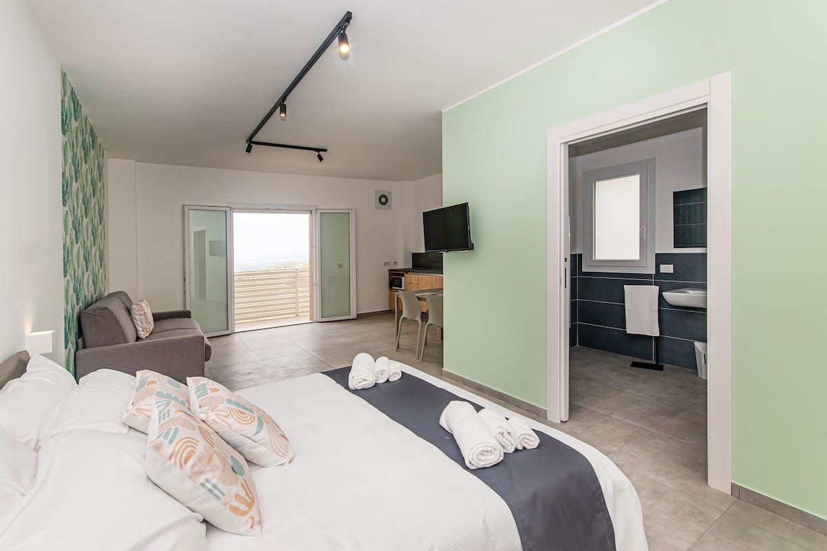 Une chambre d’hôtel moderne, équipée d’un lit, d’un canapé, d’un bureau et d’une salle de bain visible à travers une porte ouverte.