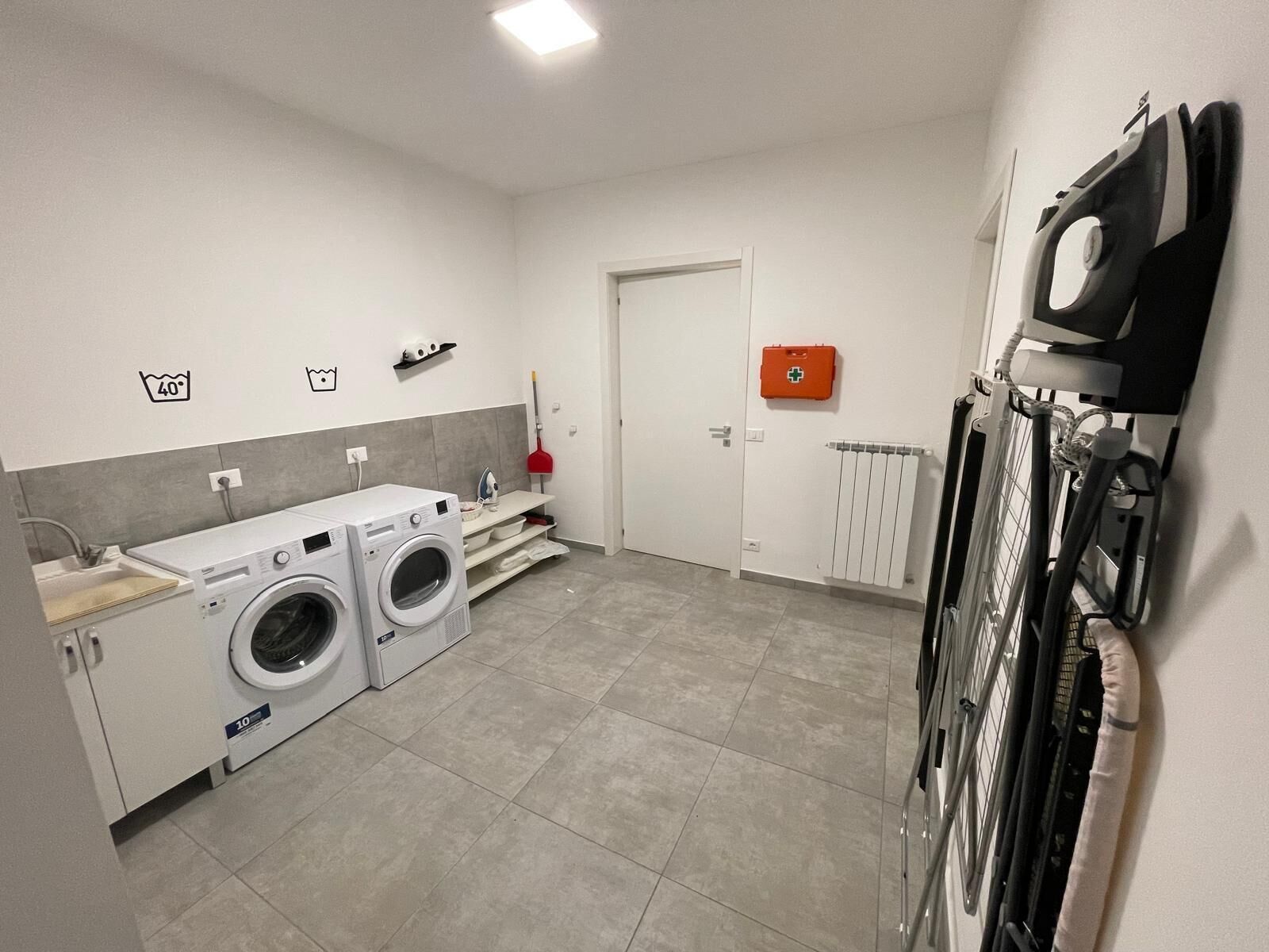 A laundry room with two washing machines, a drying rack, a radiator, and a first aid kit on the wall.