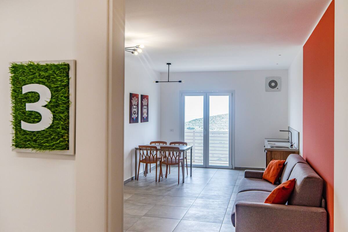Un salon moderne avec un canapé, une table à manger et des chaises, une grande fenêtre et un panneau mural portant le chiffre « 3 ».