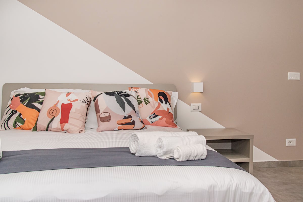 Un lit bien fait, agrémenté d’oreillers colorés, une table de chevet et une lampe fixée au mur.