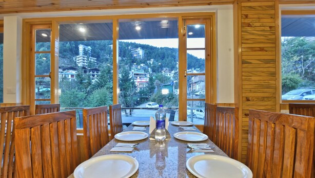 A dining area with a long table set for four, wooden chairs, and large windows offering a view of a mountainous landscape. - Hotel Sky Bhagsu (Dharamshala)