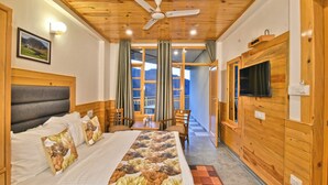 A hotel room with a bed, a television, a ceiling fan, and a view of the mountains through the windows. - Hotel Sky Bhagsu (Dharamshala)