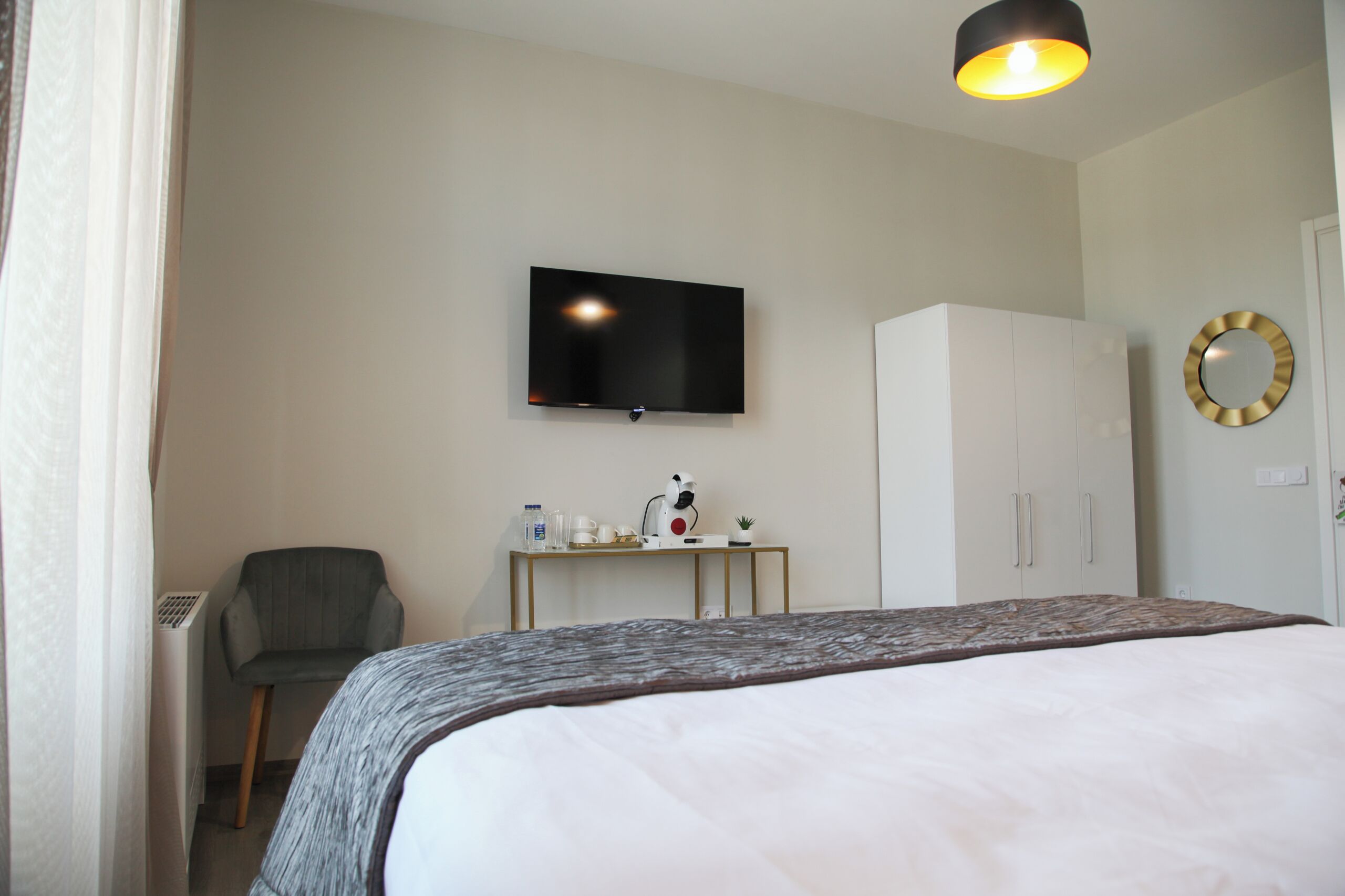 A hotel room with a bed, a desk with a flat-screen TV, a chair, a wardrobe, and a mirror on the wall.