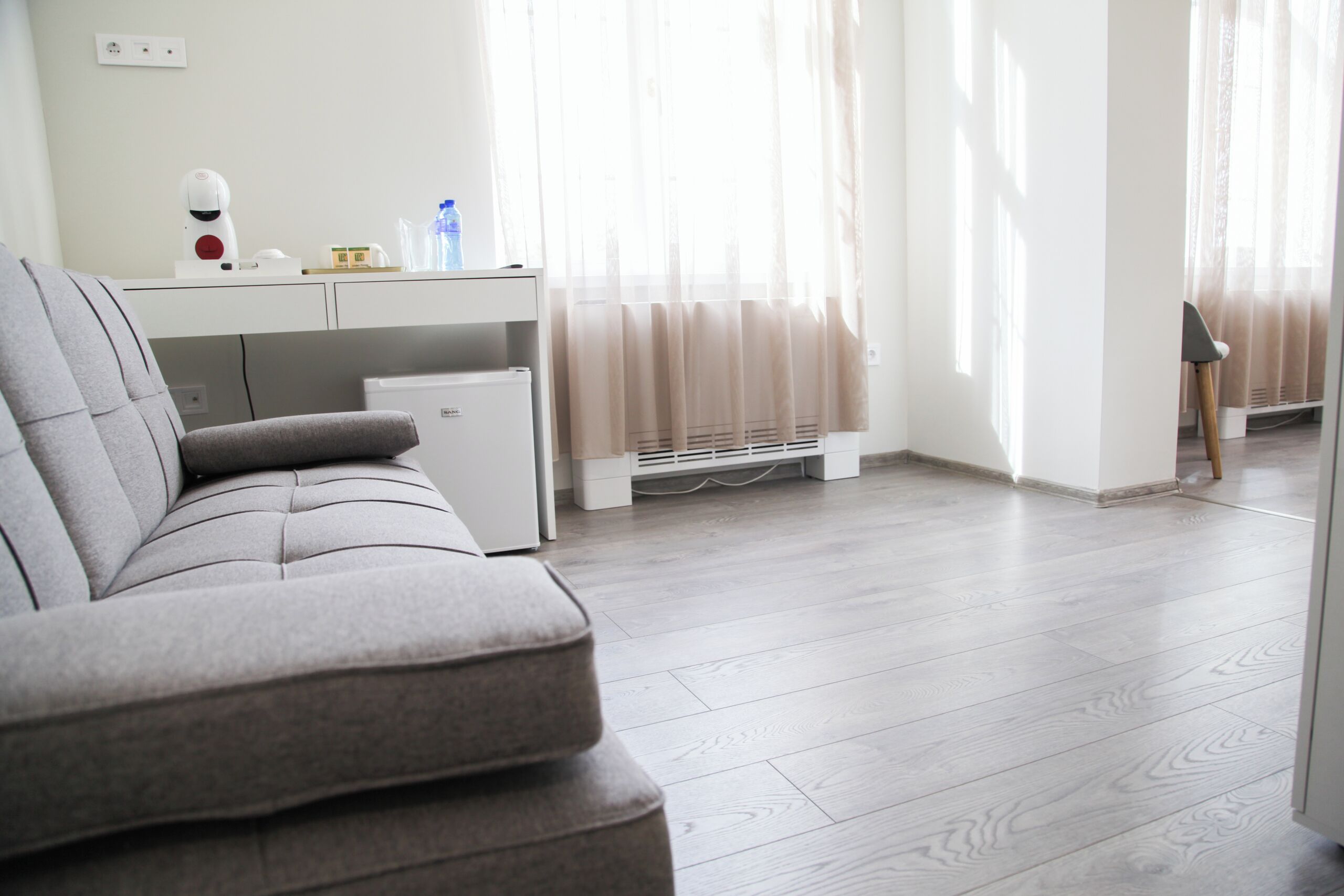 A modern living room with a grey sofa, a small white desk with a laptop, and a standing heater.