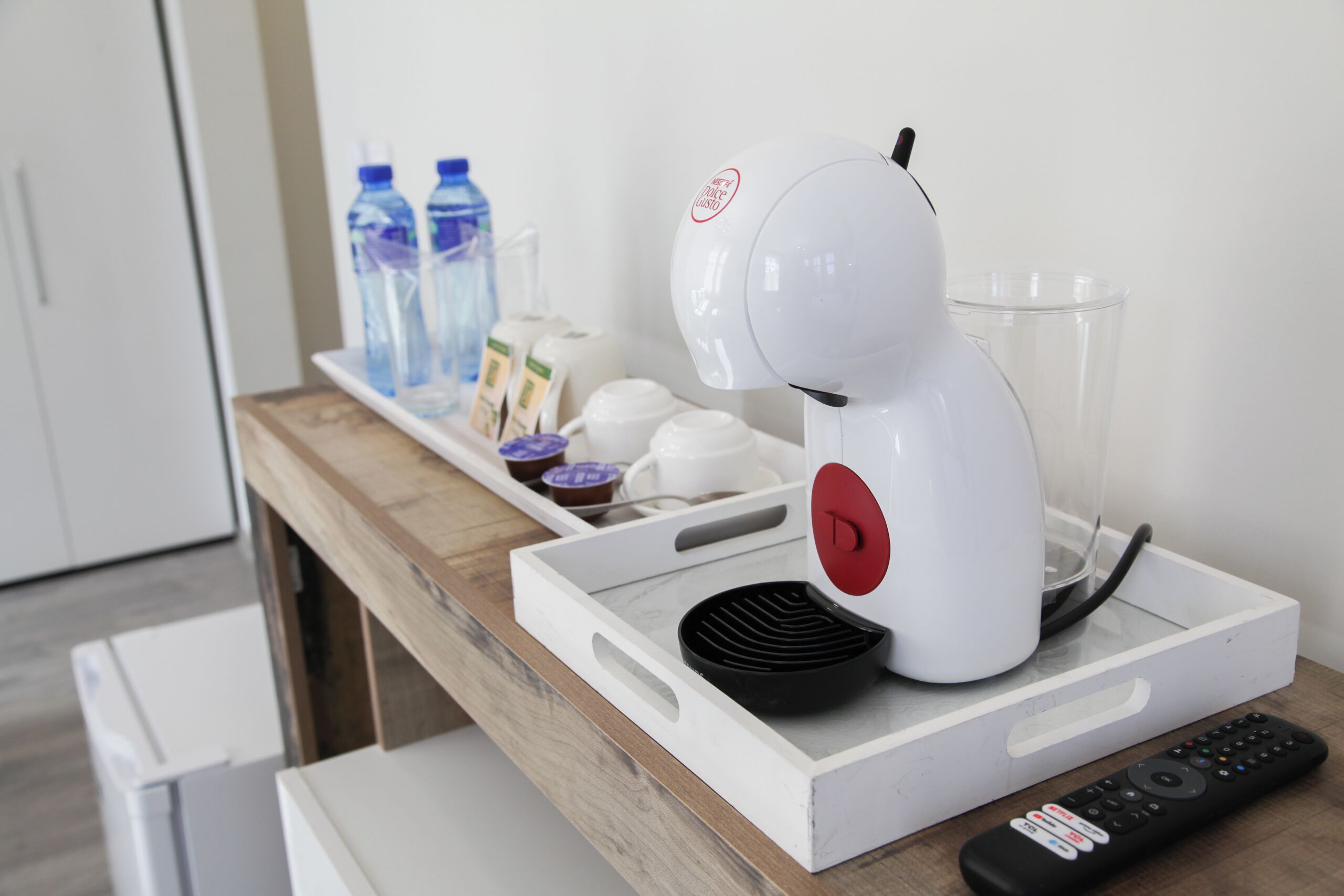 A coffee maker on a tray with a remote control, water bottles, and a glass pitcher on a wooden countertop.