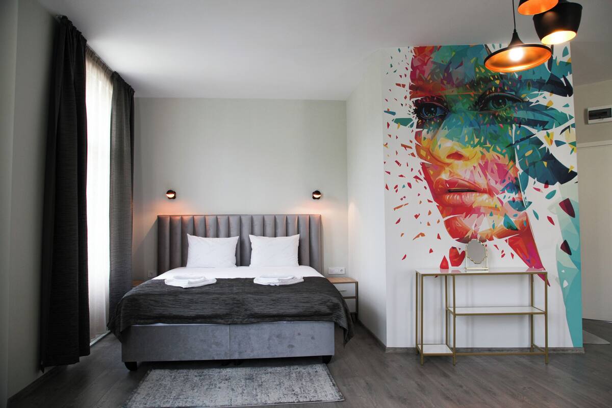 A modern bedroom with a large bed, a bedside table, a nightstand, a floor lamp, and a colorful abstract painting on the wall.