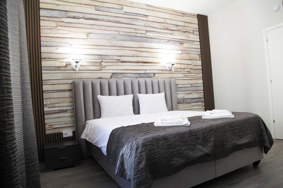 A modern bedroom with a wooden wall panel, a grey upholstered headboard, white bedding, and a small bedside table.