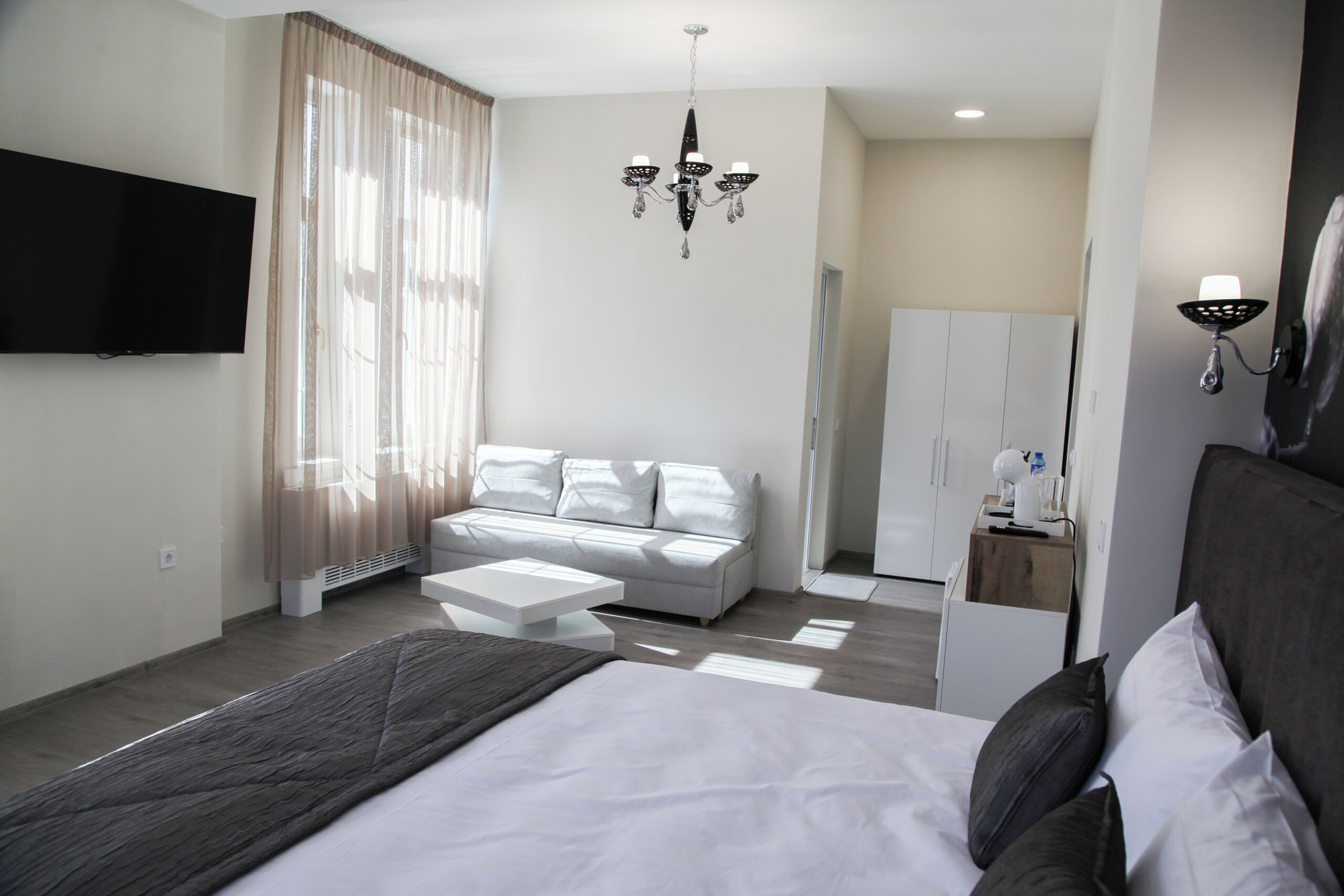 A modern hotel room with a large bed, a sofa, a TV, a chandelier, and a wardrobe.