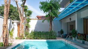 Una piscina con agua azul cristalina, rodeada de árboles y plantas, y un edificio con techo azul al fondo.