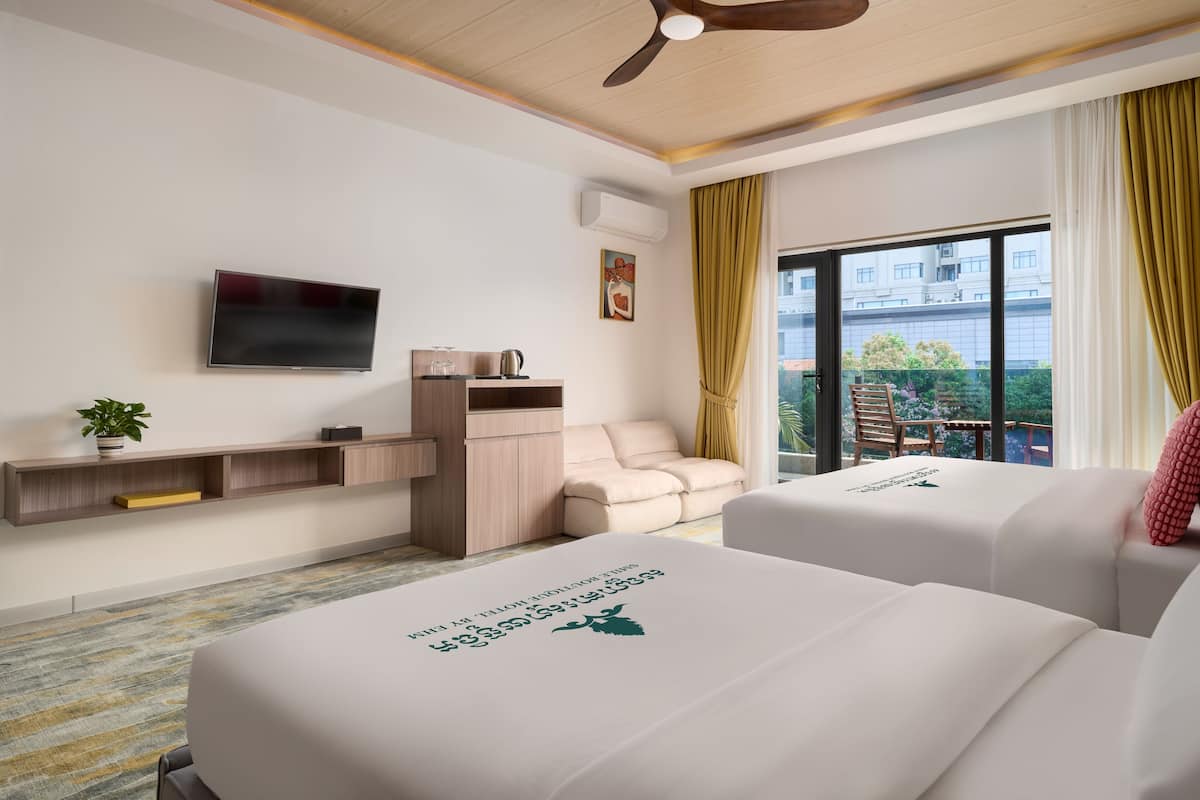 Une chambre d’hôtel avec deux lits, une télévision, un ventilateur de plafond, une fenêtre avec des rideaux, une petite table avec une plante et un canapé.
