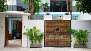 Entrada de hotel con un letrero de madera, plantas en macetas y un edificio moderno al fondo.