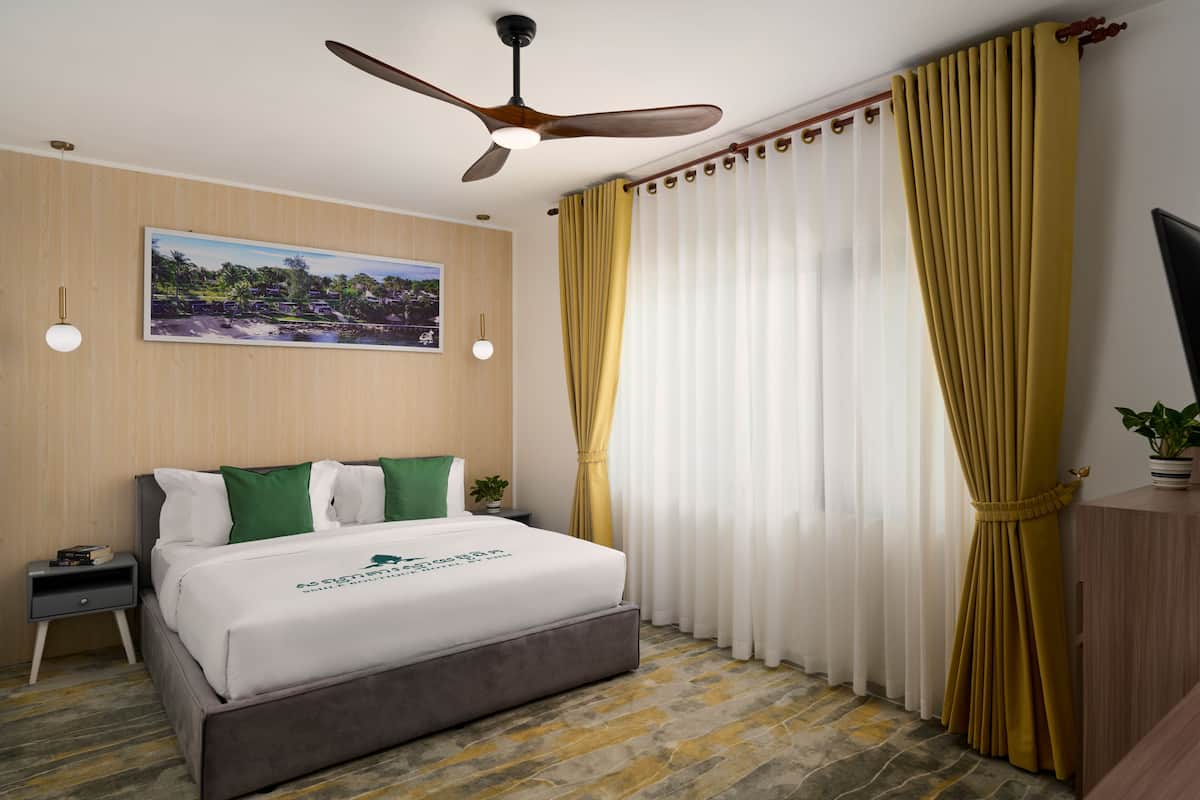 Une chambre d’hôtel avec un lit, un ventilateur de plafond, un tableau au mur et des rideaux.