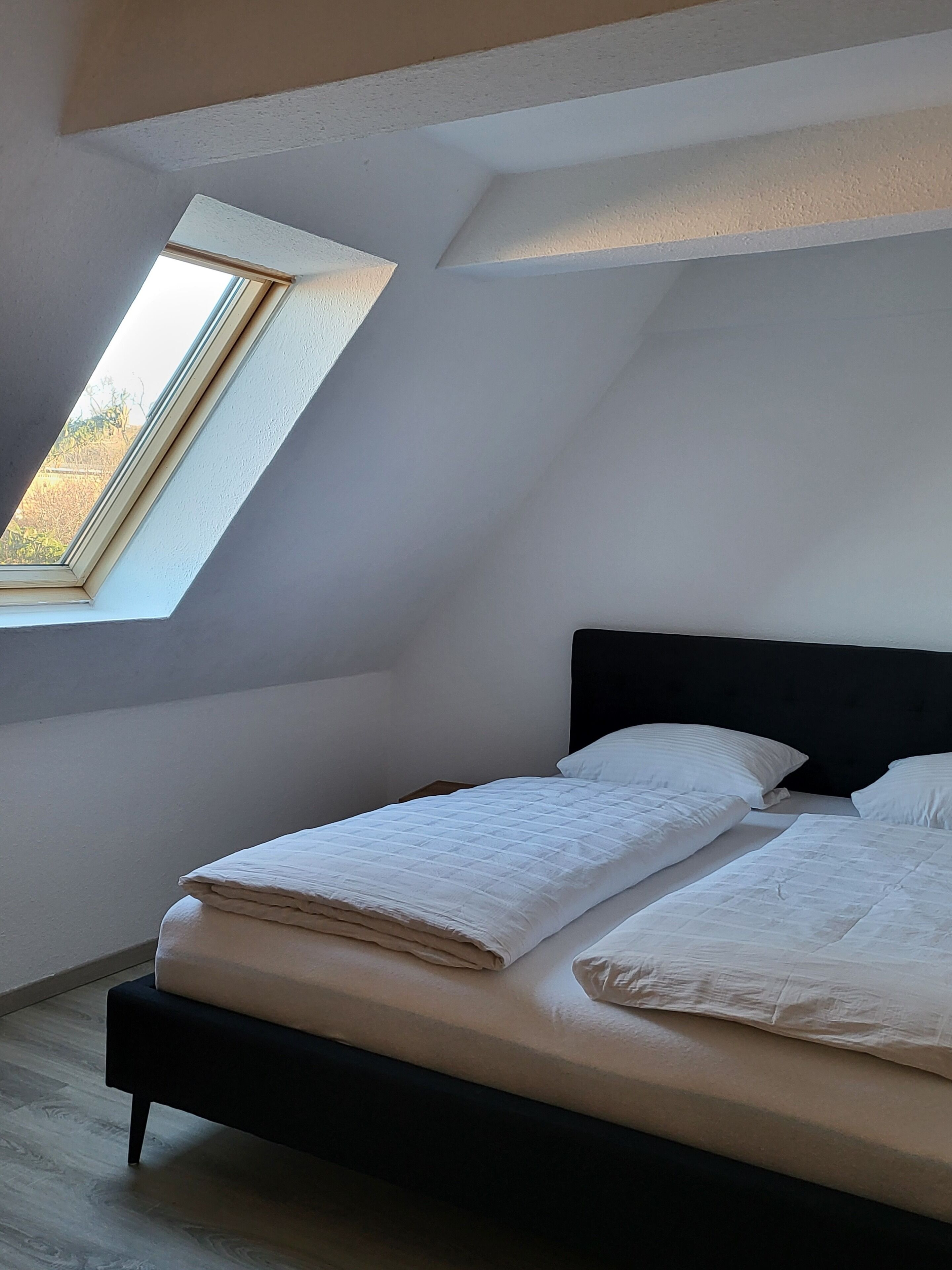 Une chambre avec une lucarne, un lit avec une literie blanche et une tête de lit sombre.