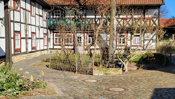 Une cour dallée de pierres, un bâtiment avec un toit recouvert de tuiles rouges et un arbre.