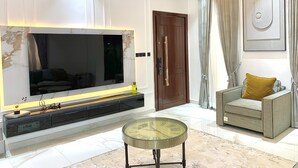 Una sala moderna con una televisión de pantalla plana grande, una mesa de centro de vidrio, un sofá con un cojín color mostaza y una puerta de madera.