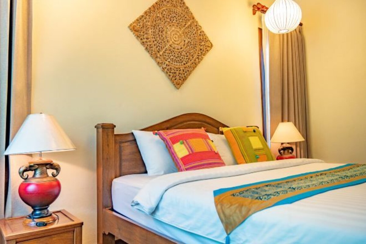 Een hotelkamer met een houten bed, een nachtkastje met lamp, een decoratief object aan de muur en een raam met gordijnen.