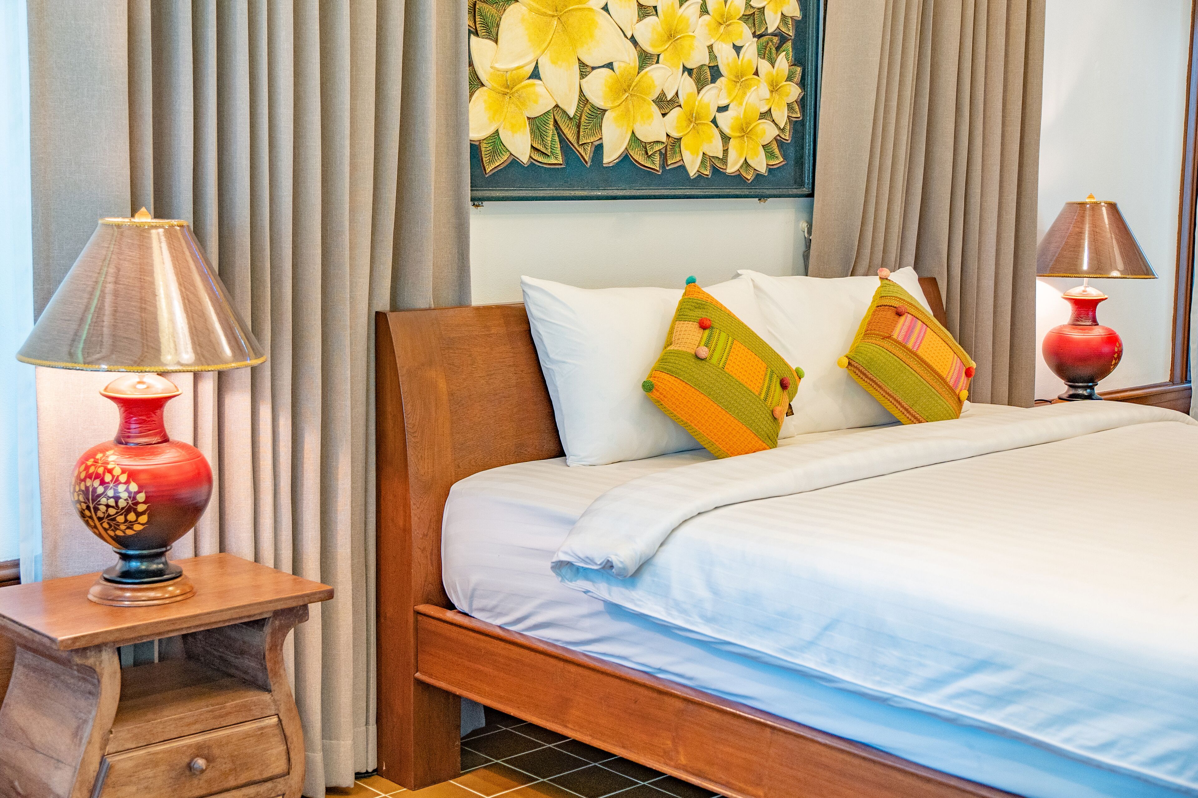 Una cama bien hecha con sábanas blancas y almohadas coloridas, una mesita de noche de madera con una lámpara roja y una obra de arte floral en la pared.