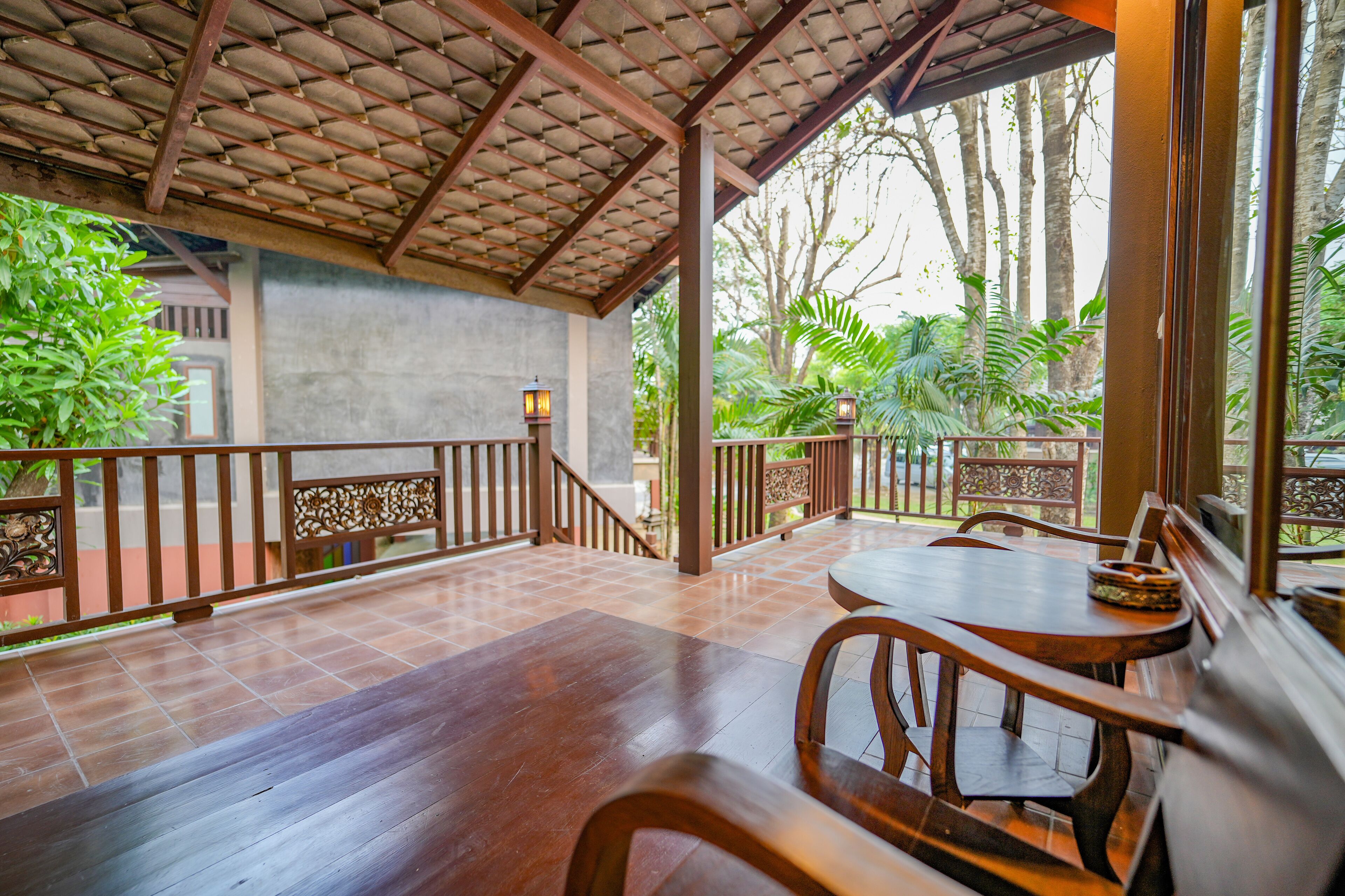 Un área de comedor al aire libre con mesas y sillas de madera, piso de baldosas y baranda con vista a un jardín con árboles y plantas.