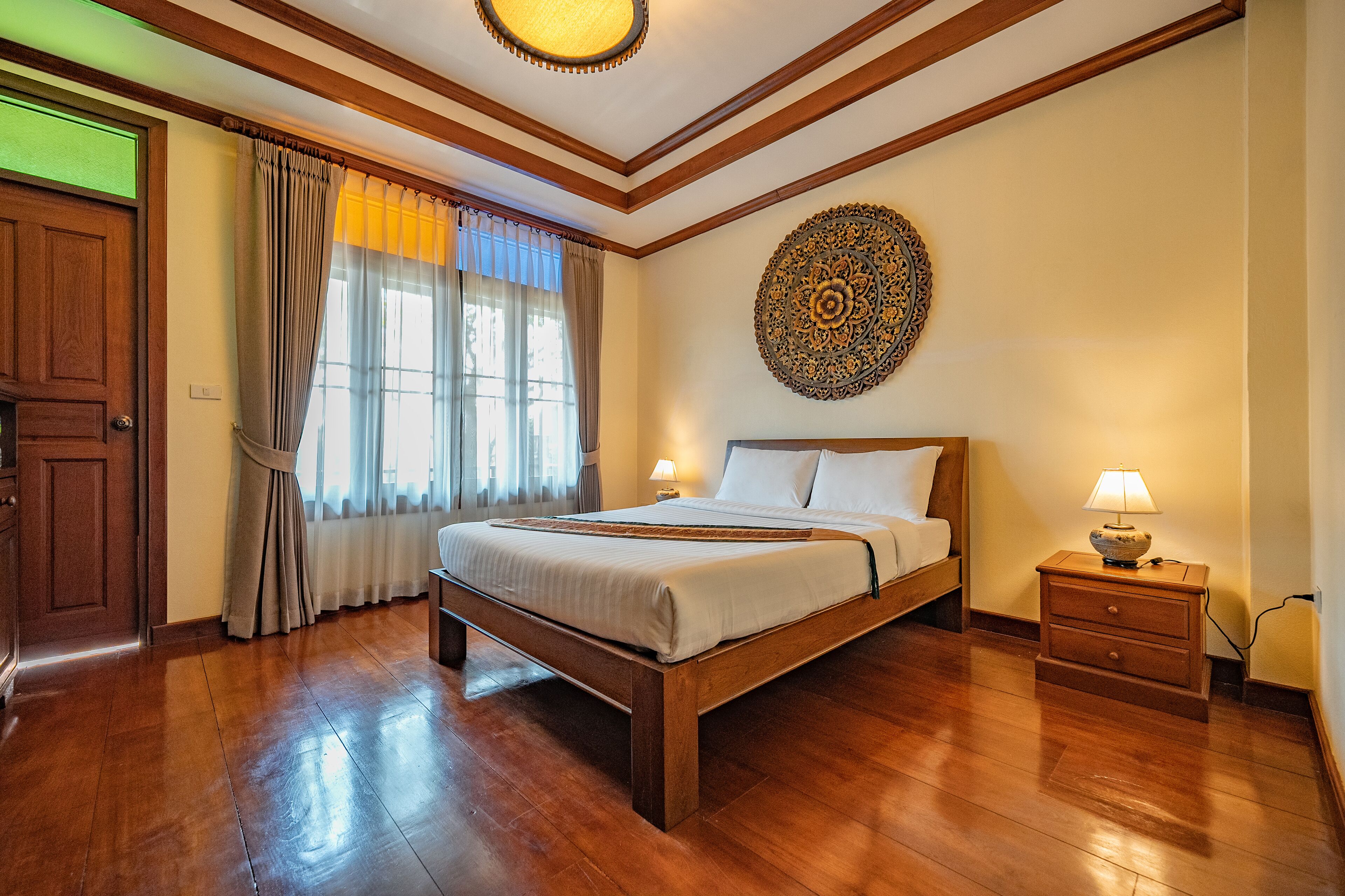 Un dormitorio con una cama de madera, una mesita de noche con lámpara, un adorno en la pared y un amplio ventanal con cortinas.