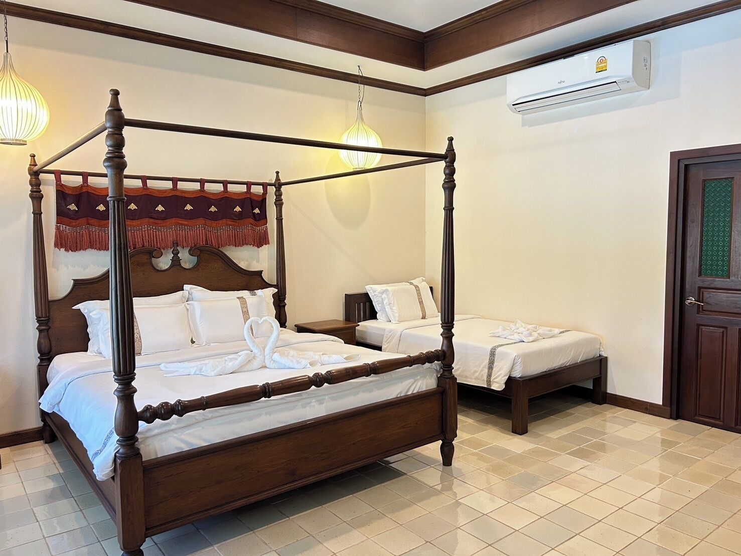 A four-poster bed with white linens and two smaller beds in a room with tiled flooring and an air conditioning unit.