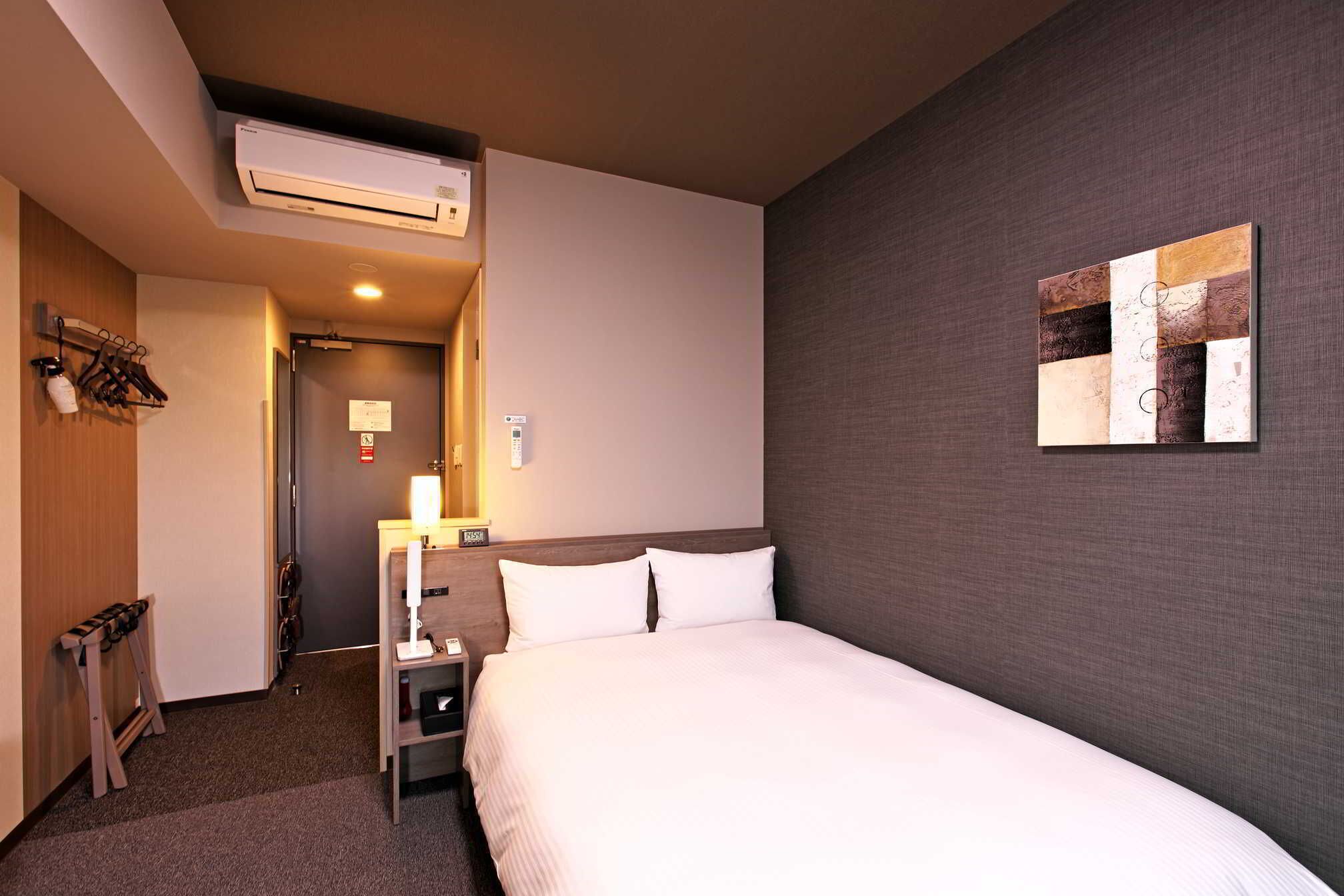 A hotel room with a bed, bedside table, lamp, and an air conditioning unit.