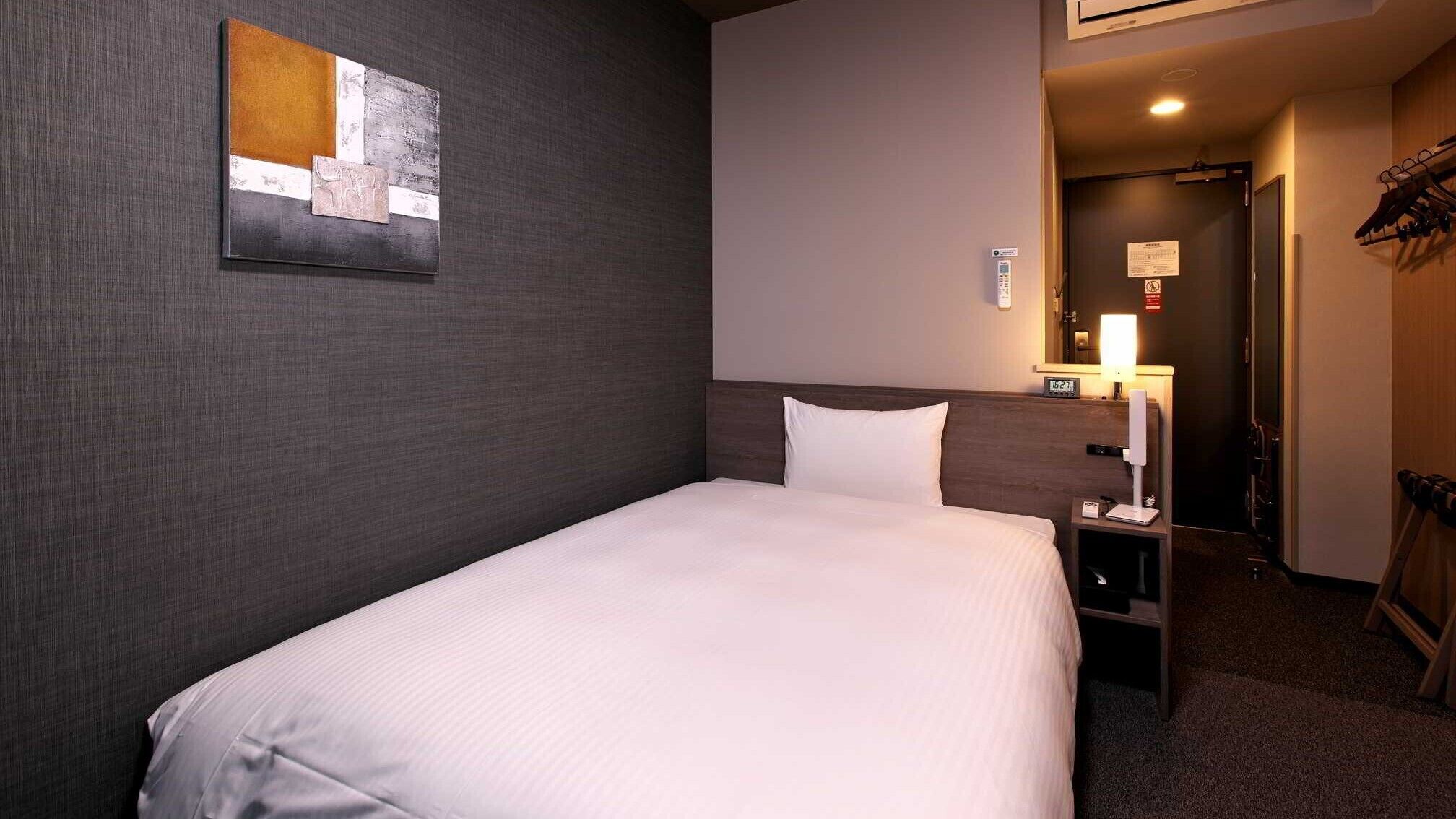 Habitación de hotel con una cama individual, una mesita de noche, una lámpara de pared, un reloj y un perchero.