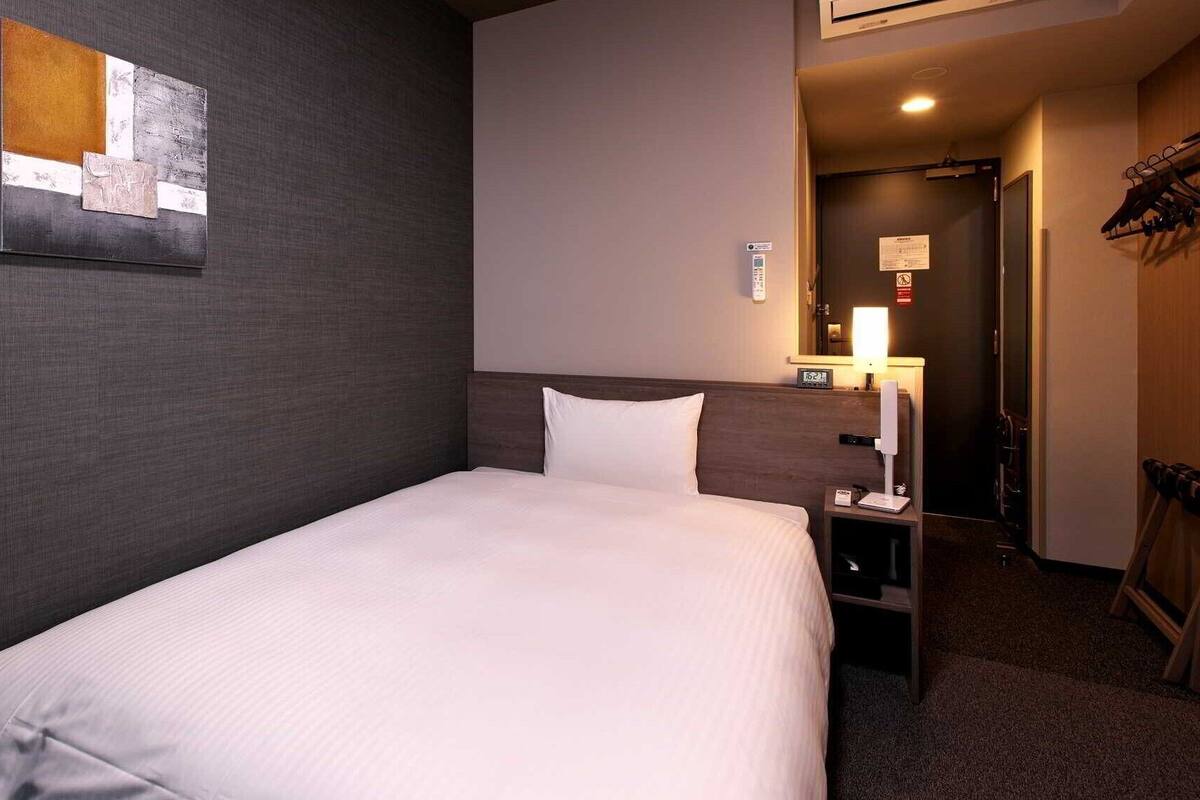 A hotel room with a single bed, a nightstand, a wall-mounted lamp, a clock, and a coat rack.