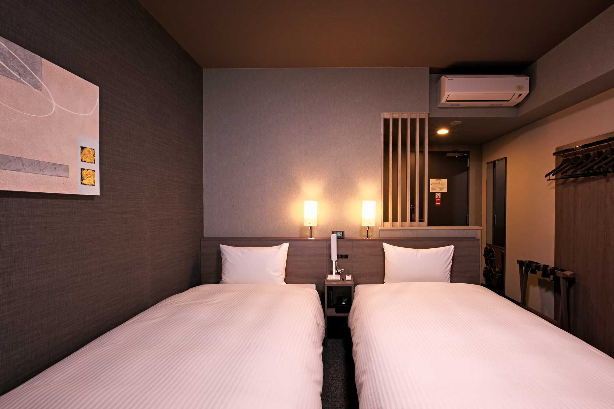 Habitación de hotel con dos camas, una mesita de noche, lámparas de pared y un equipo de aire acondicionado, además de un perchero.