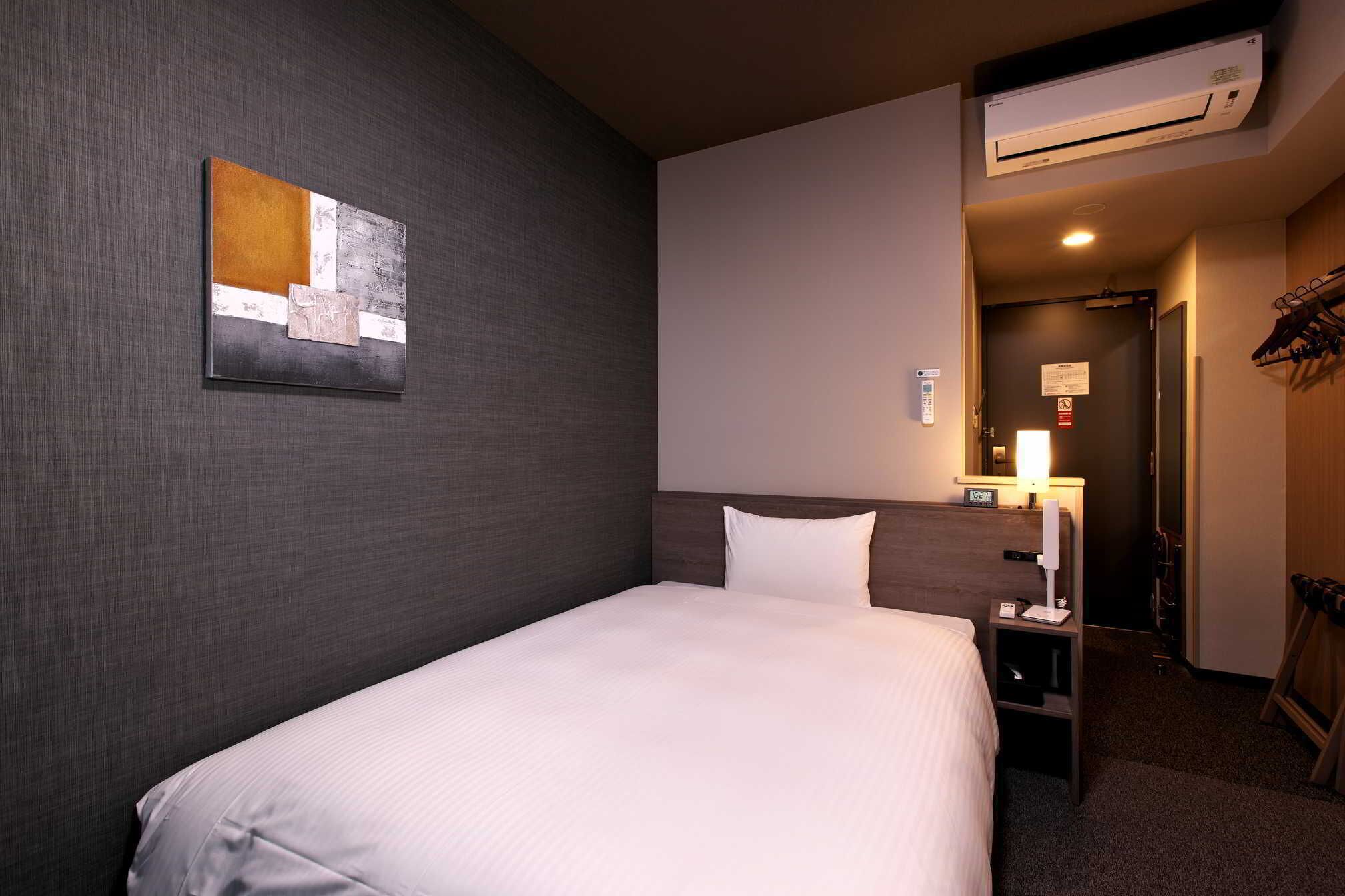 A hotel room with a single bed, a nightstand, a wall-mounted air conditioner, and a door leading to a hallway.