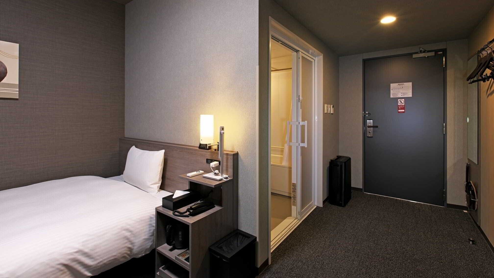 Una habitación de hotel con cama, mesita de noche, lámpara y puerta que da a la zona de ducha.