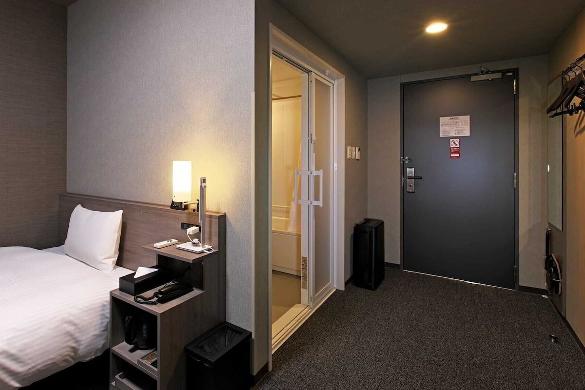 A hotel room with a bed, bedside table, lamp, and a door leading to a shower area.