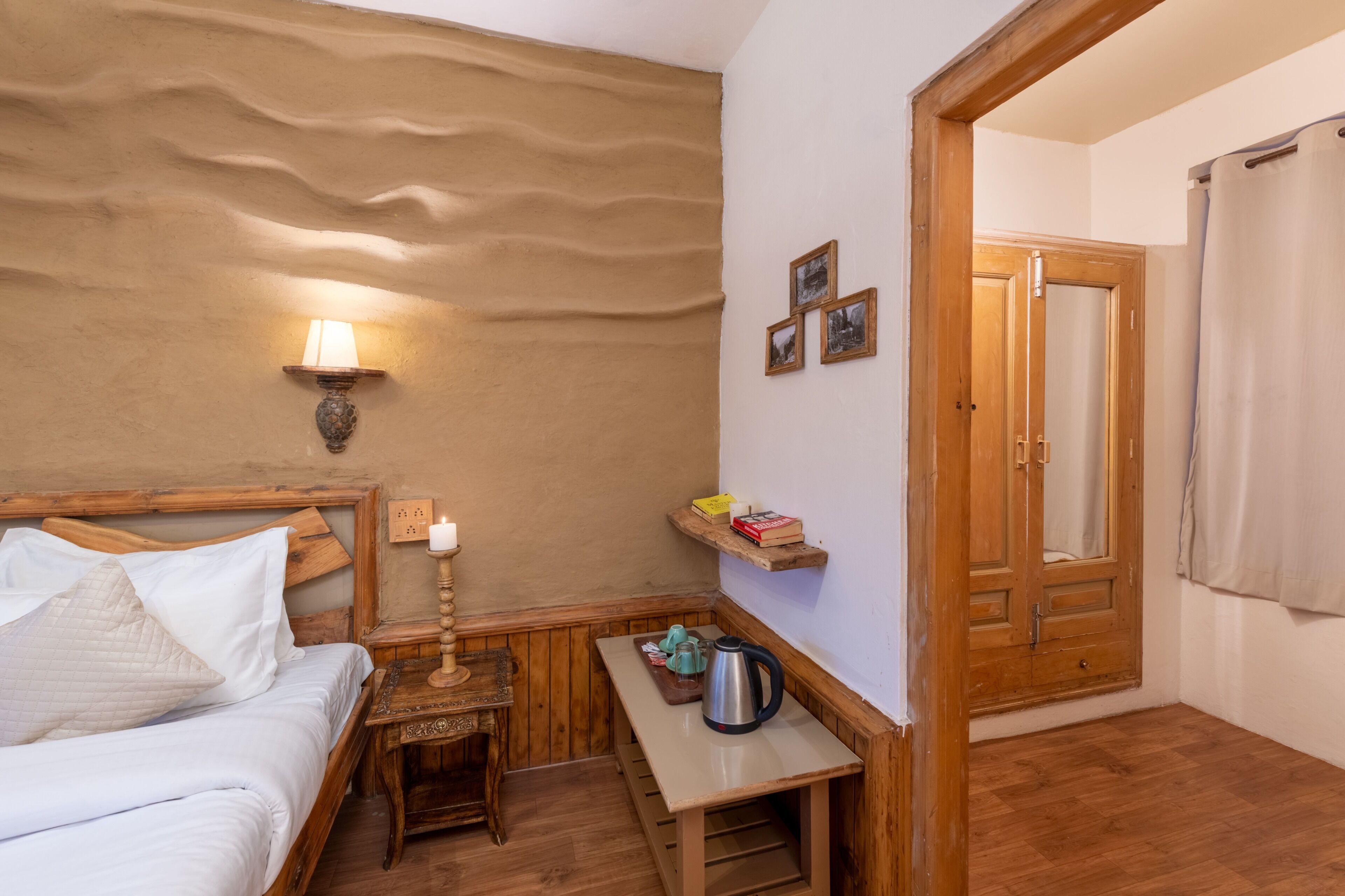 A room with a bed, a nightstand with a lamp, a small table with a kettle, and a wooden door.