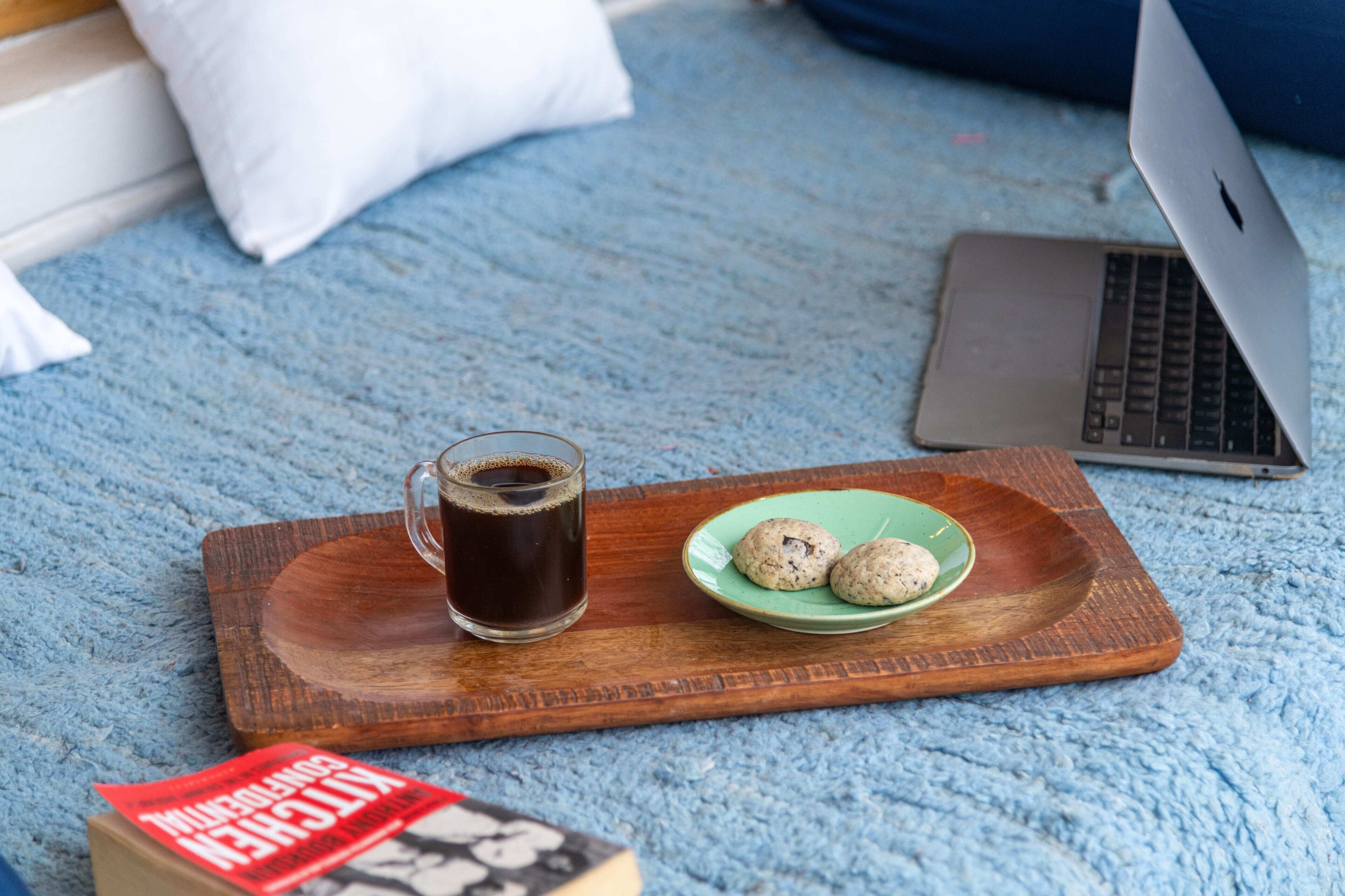 A laptop on a blue carpet, a wooden tray with a glass of coffee and cookies, and a book.
