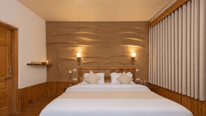 A hotel room with a wooden floor, a large bed with white bedding, a wooden headboard, two wall-mounted lamps, and a wooden door. - Beholden Stay & Cafe-Mountain View Boutique Stay in Old Manali (Manali)
