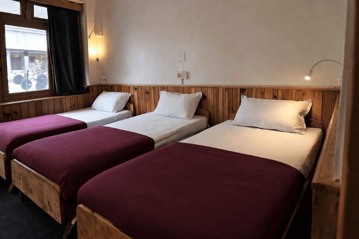 Een kamer met drie houten bedden, elk voorzien van een witte matras en een rode spreideken. Er zijn wandlampen en een raam met gordijnen.