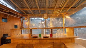 A rustic wooden bar area with stools, a counter with glasses and bottles, and a view through a large window. - Beholden Stay & Cafe-Mountain View Boutique Stay in Old Manali (Manali)