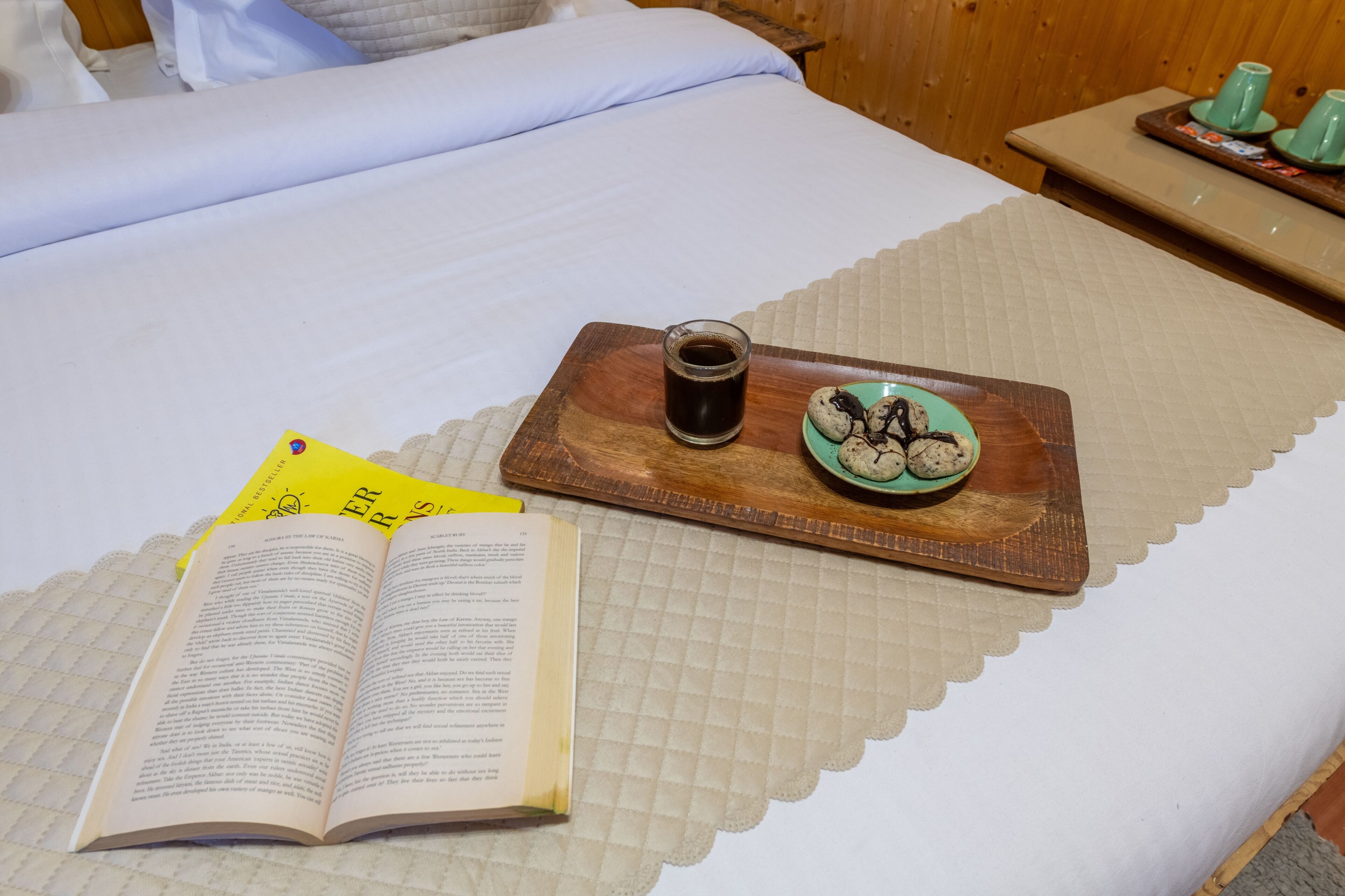 A bed with a quilted bedspread, a tray with a mug of coffee and cookies, and an open book.