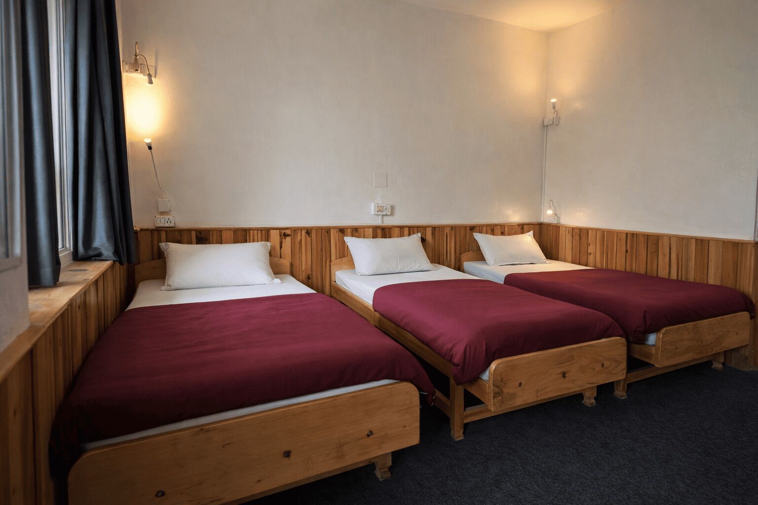 Um quarto com três camas de madeira, cada uma com colcha vermelha e almofadas brancas, e uma janela com cortinas.