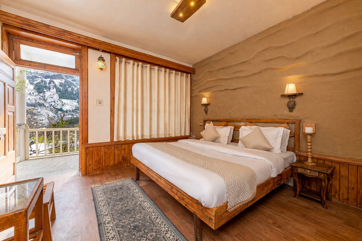 A bedroom with a wooden bed, a balcony with a view of snow-covered mountains, and a small table with chairs.