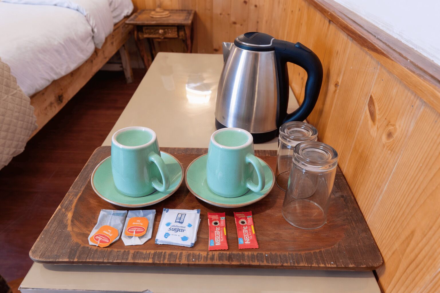 A wooden table with two green cups, a kettle, and packets of tea and sugar.