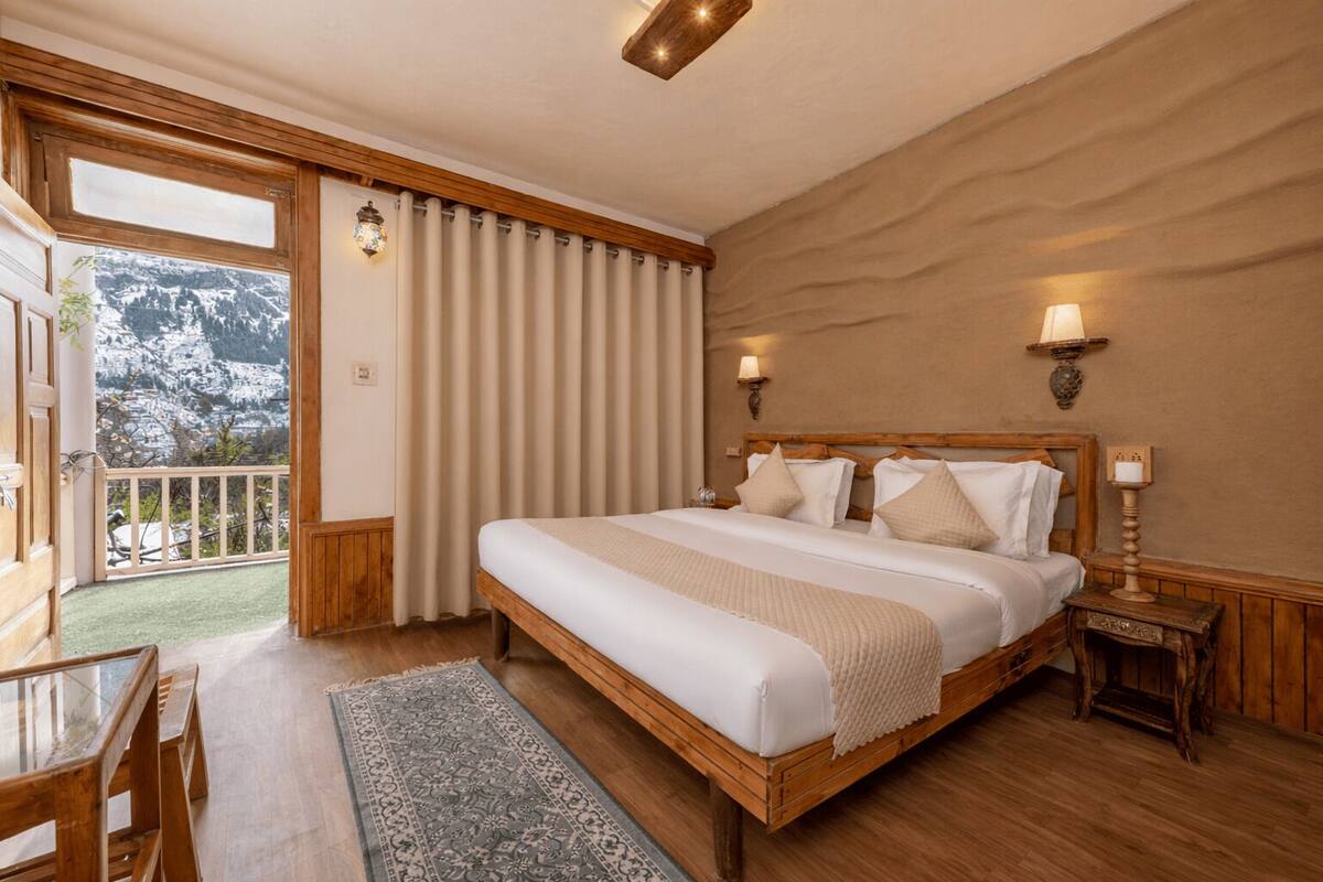 Een slaapkamer met een groot bed, een houten hoofdbord, twee bedlampen, een nachtkastje, uitzicht op bergen door een schuifdeur en een balkon met een tafel en stoelen.