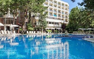 Piscina con sillas de descanso y sombrillas, rodeada de árboles y un edificio de varios pisos.