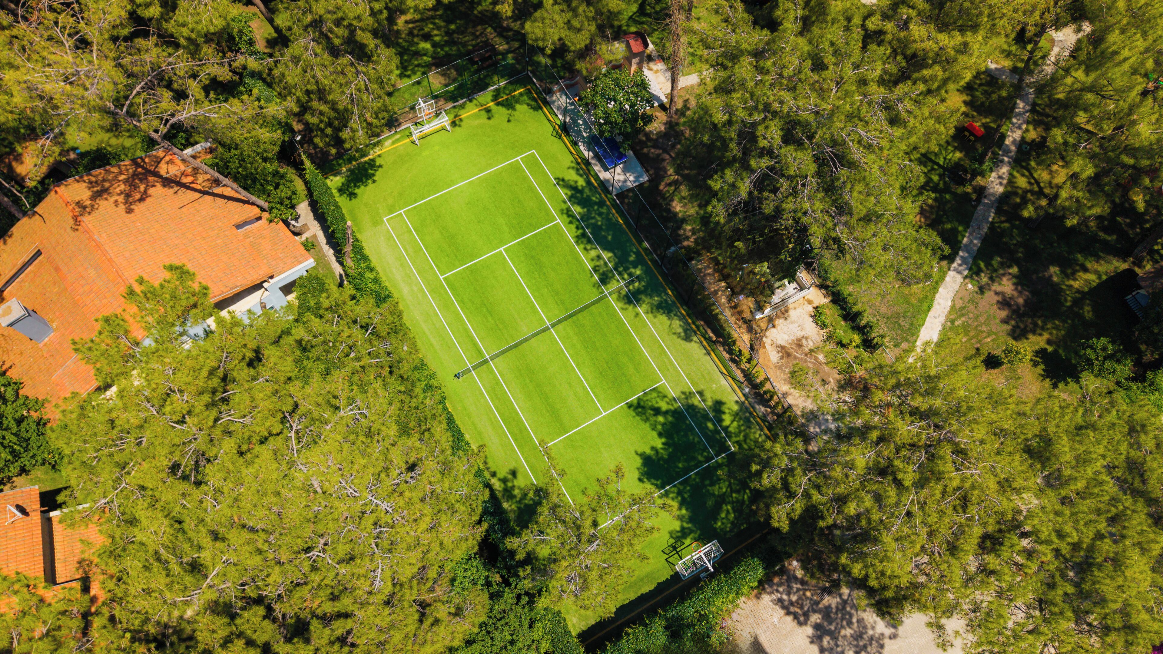 Vista aérea de una cancha de tenis rodeada de árboles, con un edificio a la izquierda.
