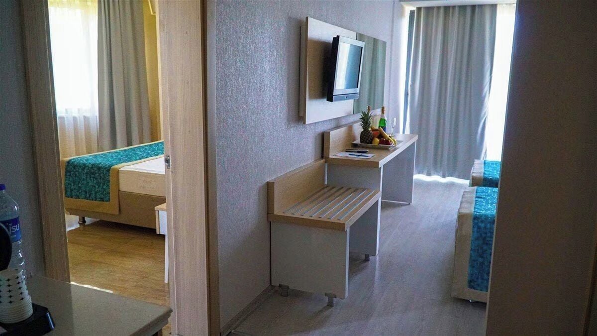 Habitación de hotel con una cama, un escritorio, un televisor y una pequeña mesa con frutas y una botella.