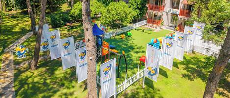 A park with playground equipment, a white picket fence, and a building with a red balcony.
