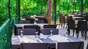 Un área de comedor al aire libre con mesas y sillas de mimbre, rodeada de abundante vegetación.