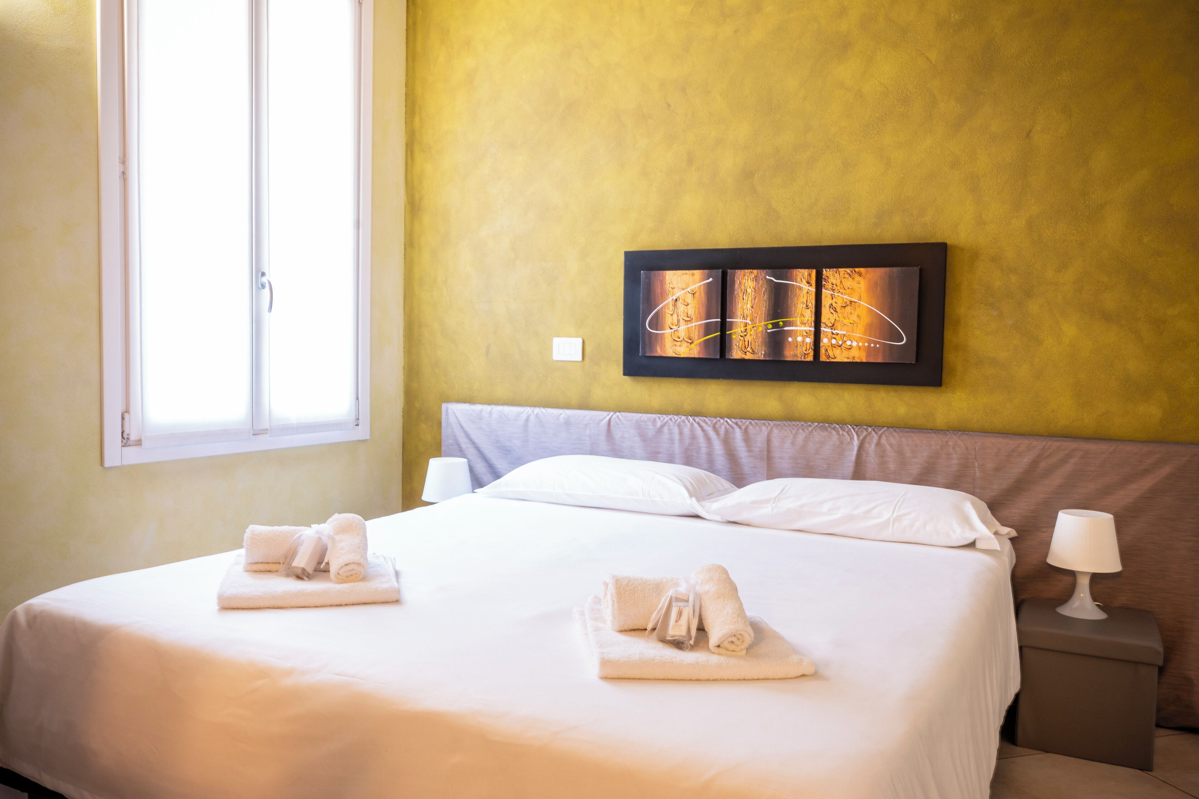 A hotel room with a large bed, two pillows, a nightstand with a lamp, and a wall art piece.