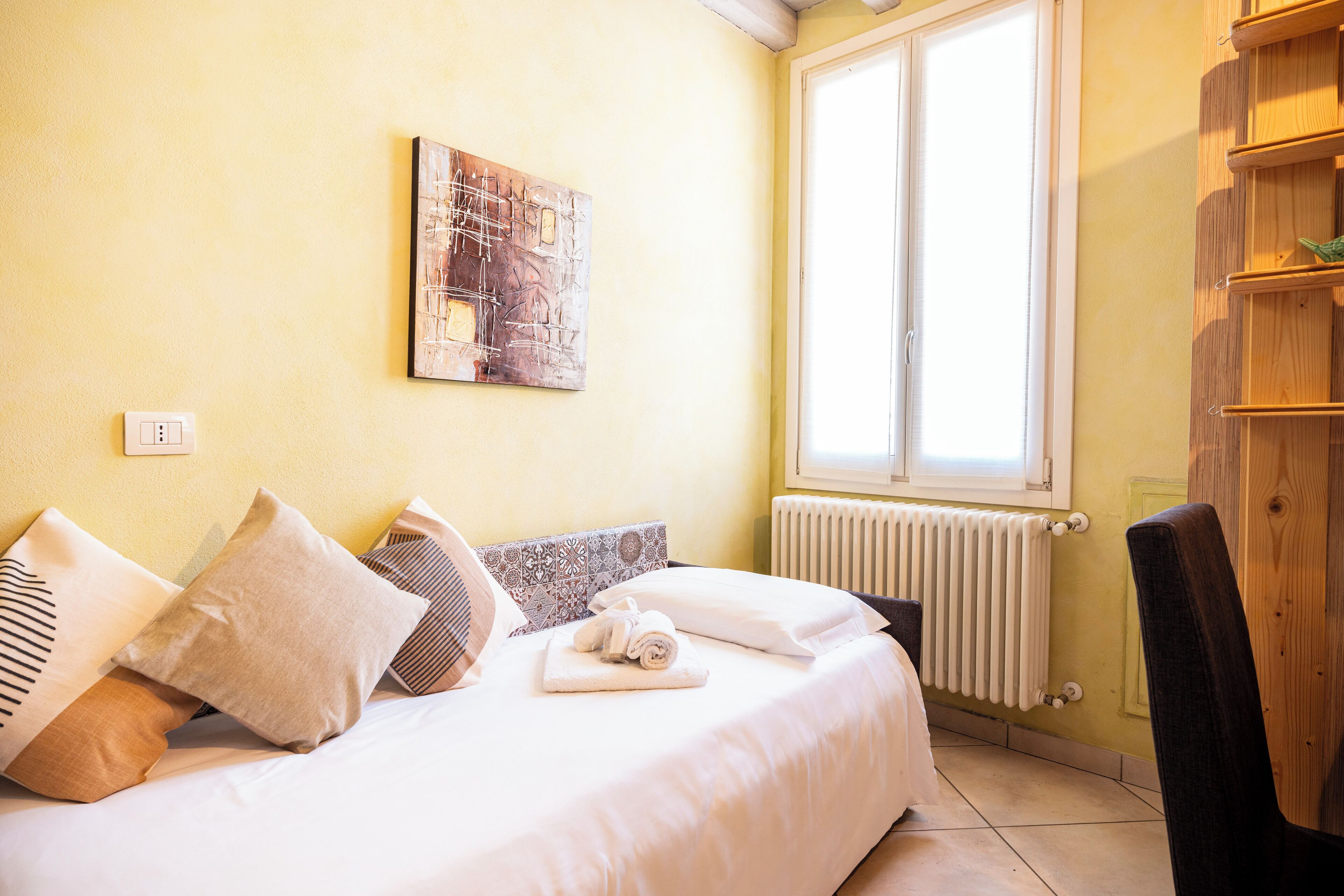 A bedroom with a bed, pillows, a rolled towel, a radiator, a window, and a wooden chair.