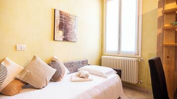 A bedroom with a bed, pillows, a rolled towel, a radiator, a window, and a wooden chair.